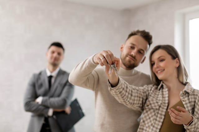 Satisfied couple holding a set of keys with an agent standing behind them and watching proudly